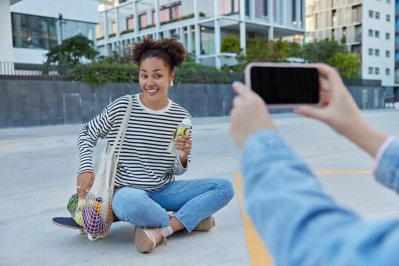 Happy teenage girl poses for a smartphone photo, sitting cross-legged on a skateboard, carrying a net bag with fruits and holding a fresh citrus detox drink, spending her free time in an urban place.