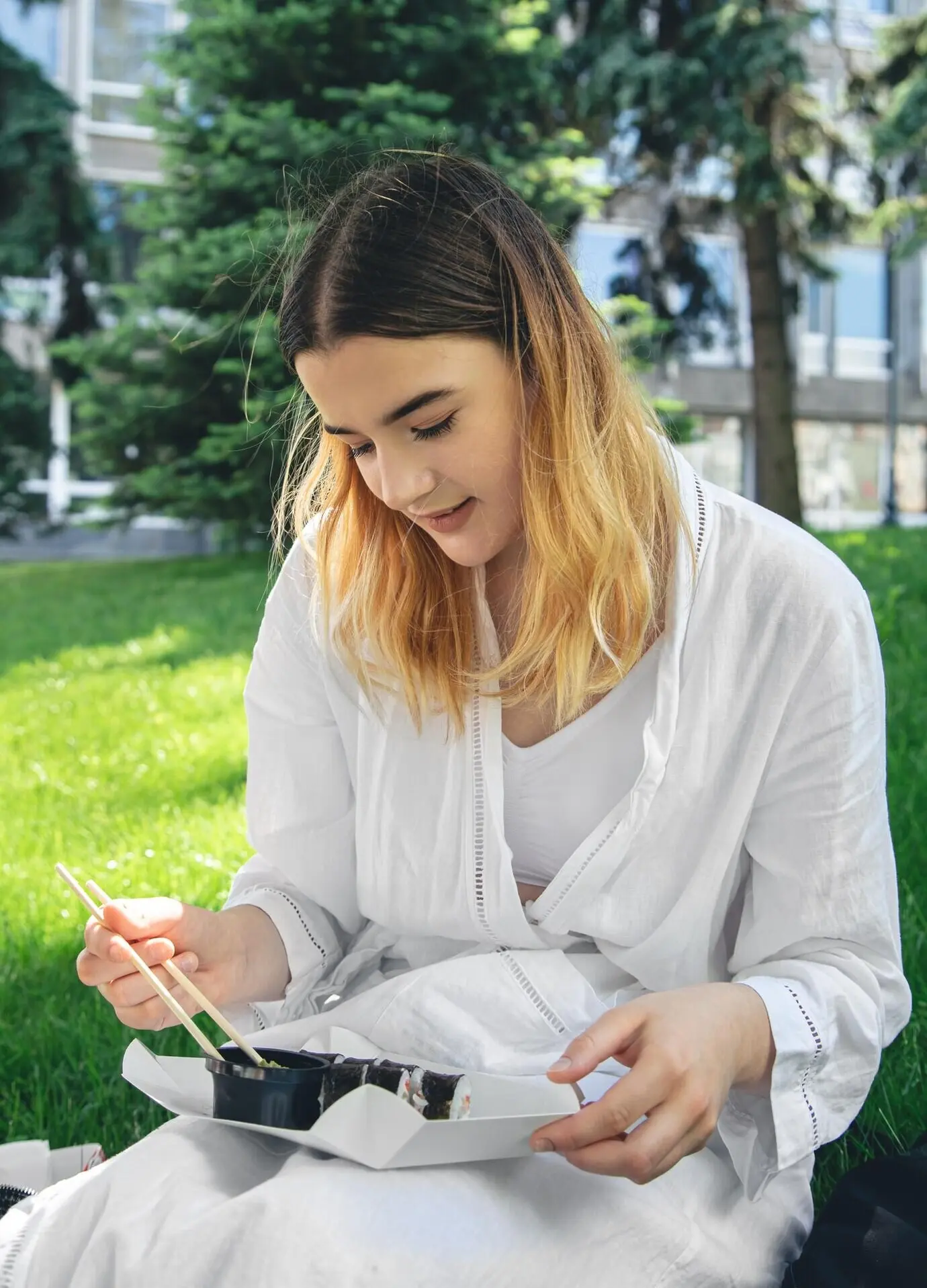 A beautiful girl is sitting on the grass in the park, eating sushi.