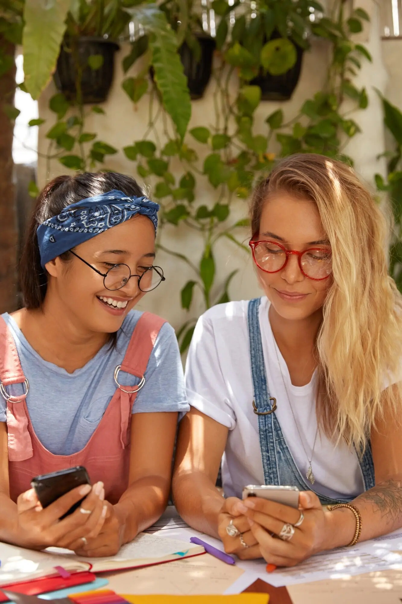 A shot of women of diverse ethnicities who are sharing multimedia files via Bluetooth, holding modern smartphones, sitting at a desk, collaborating on a common task, wearing transparent glasses, and addicted to modern technologies.